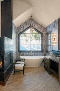 Snowy fields out the window of a freestanding bathtub in a large elegant bathroom with cozy corner fire.